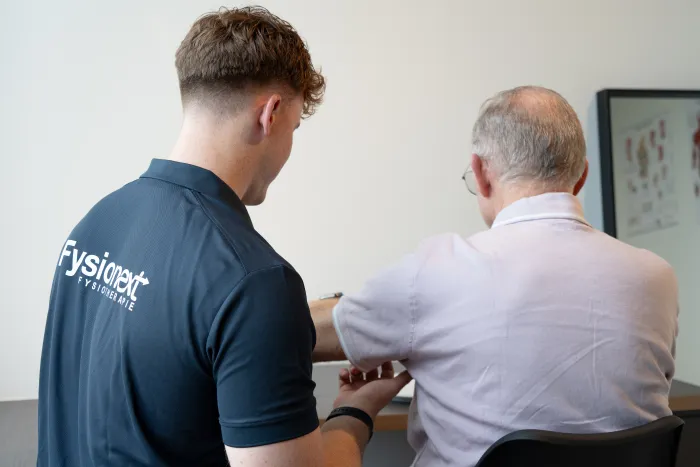 Fysiotherapeut in FysioNext-poloshirt bespreekt de behandeling met een oudere patiënt