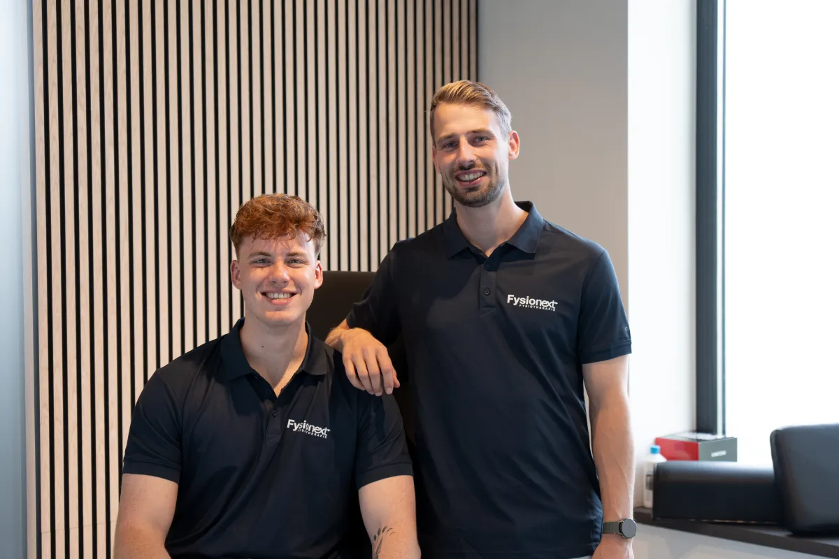 Twee fysiotherapeuten van het FysioNext-team in een FysioNext-poloshirt poseren samen in de praktijk